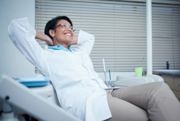 Relaxed,Smiling,Female,Dentist,Sitting,On,Chair,With,Hands,Behind